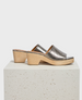 A pair of open-toe mule sandals featuring a metallic silver leather upper with a textured finish, set on a light wooden block heel and platform with a smooth surface. 1