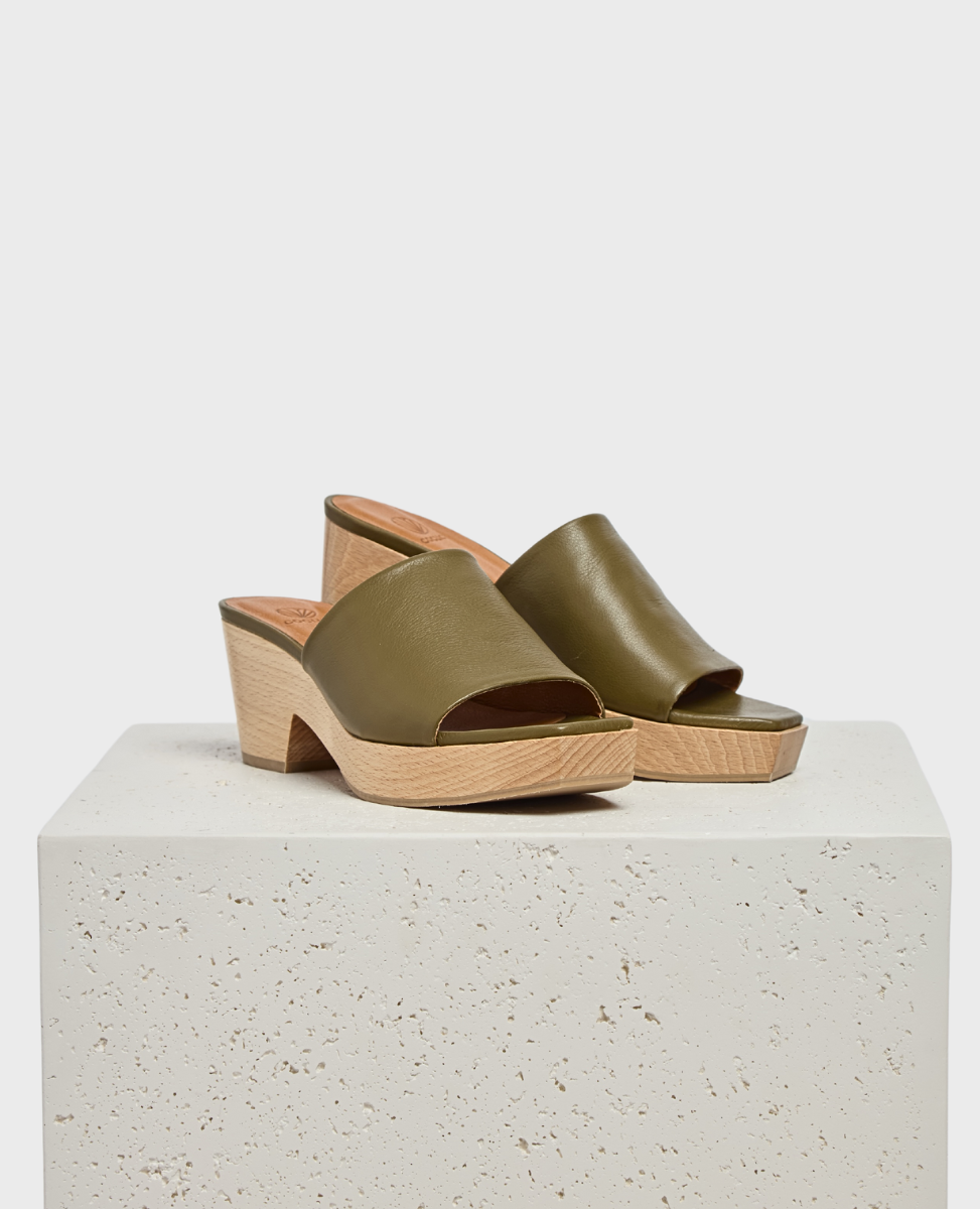 Olive green leather slide sandals feature a smooth upper with an open toe design, set on a light wooden block heel and platform with a textured finish.