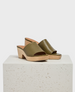 Olive green leather slide sandals feature a smooth upper with an open toe design, set on a light wooden block heel and platform with a textured finish. 1