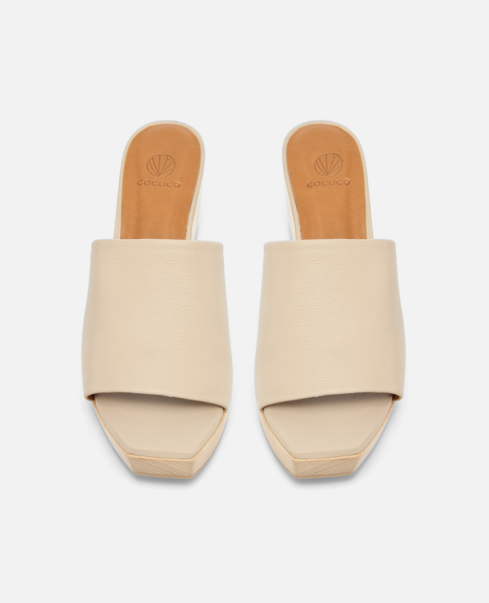 Beige slip-on sandals with a wide, smooth leather strap, open toe design, and a flat, square-shaped sole featuring a subtle logo imprint on the insole.