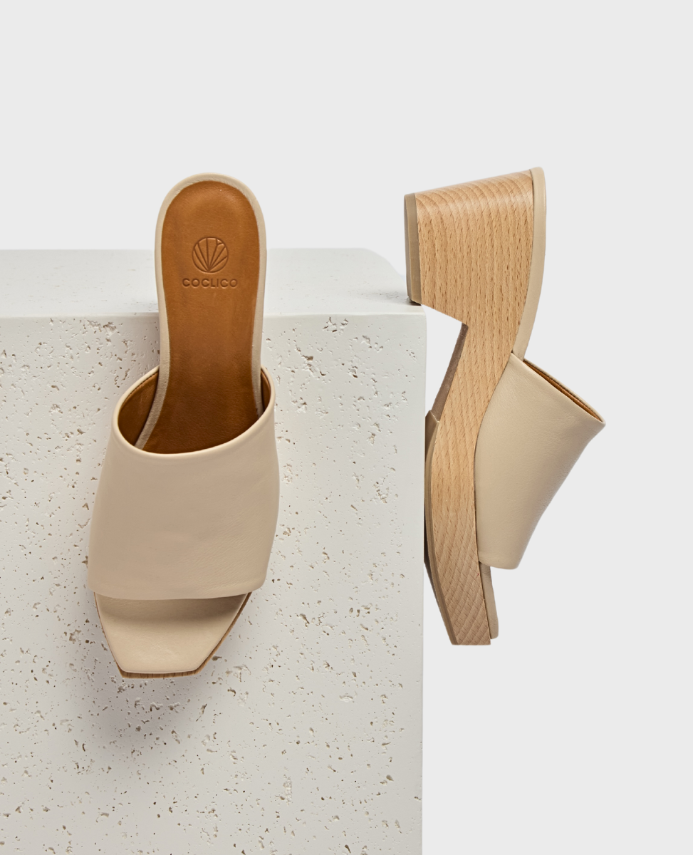 A pair of beige, open-toe mules with a smooth leather upper, featuring a chunky wooden block heel and a minimalist design.