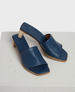 A pair of elegant blue leather slide sandals with a square toe, featuring a low wooden block heel and a smooth, minimalist design. 2