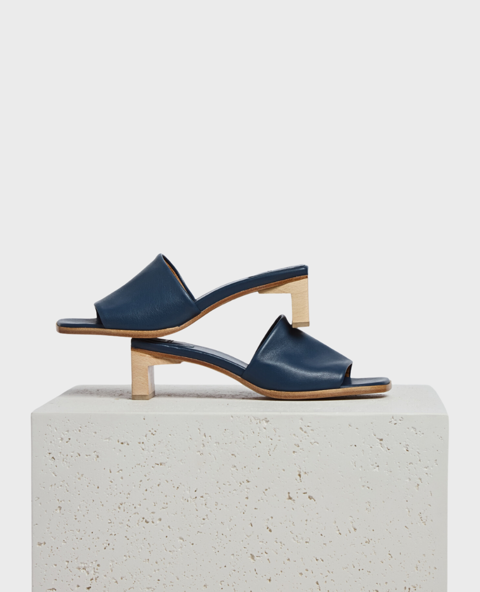 A pair of sleek, navy blue leather sandals with a minimalist design featuring a wide strap across the foot, an open toe, and a unique wooden block heel, displayed on a textured, light-colored stone surface.