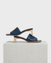 A pair of sleek, navy blue leather sandals with a minimalist design featuring a wide strap across the foot, an open toe, and a unique wooden block heel, displayed on a textured, light-colored stone surface. 1