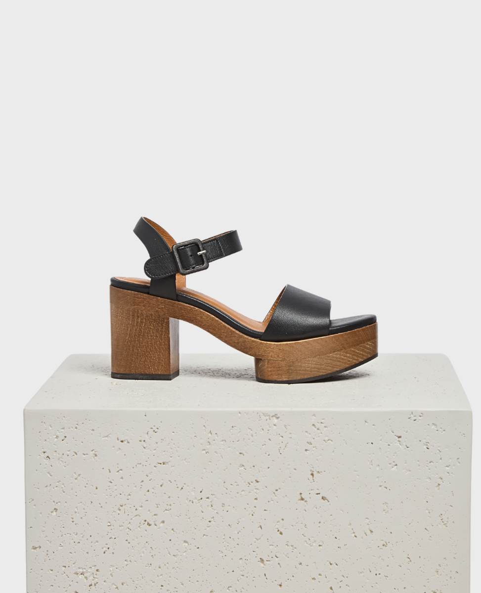 A black leather sandal with a thick strap across the toes and an adjustable ankle strap, featuring a chunky wooden platform and heel, displayed on a textured white surface.