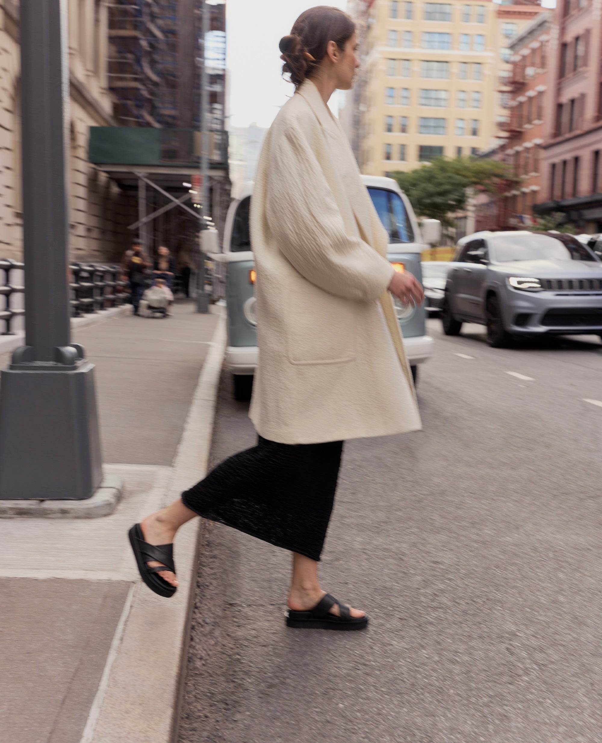 Woman in a beige coat and footbed sandals walking on a city street