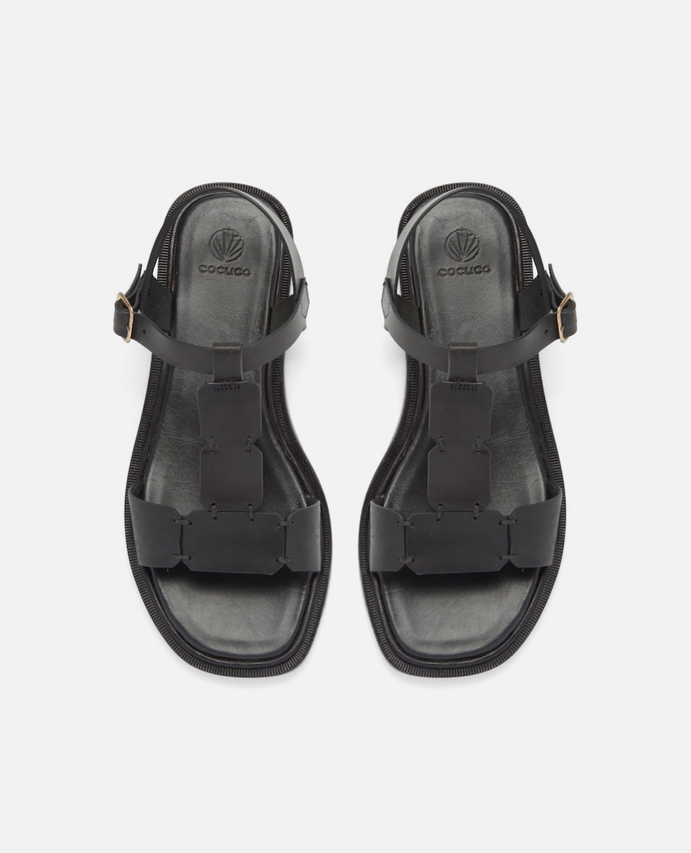 Top view of a black leather T-strap sandal with hand-linked panels, slim ankle strap, and a sleek flexible leather sole with a textured welt.