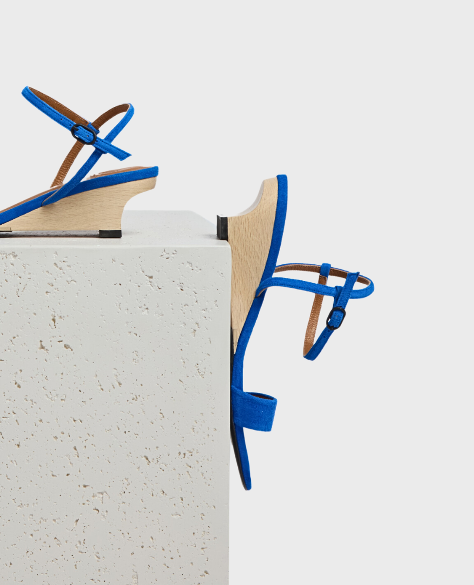 A pair of vibrant blue sandals with a minimalist design featuring thin straps and a wooden wedge heel, displayed on a textured white surface.