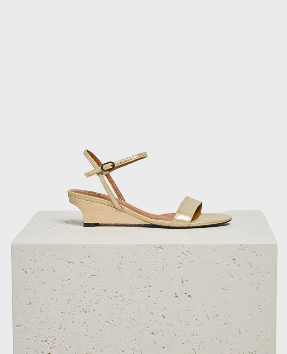 A gold sandal with a low wedge heel, featuring a sleek metallic finish, an open toe design, a thin ankle strap with a small buckle, and a smooth, slightly textured sole.