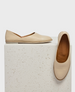 A pair of sleek, beige leather flats with a smooth texture, featuring a minimalist design and a slightly pointed toe, displayed on a textured white surface. 1