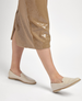 A pair of sleek, beige slip-on shoes with a smooth leather texture, featuring a rounded toe and a flat sole, worn by a person in a shimmering, light brown skirt. 4