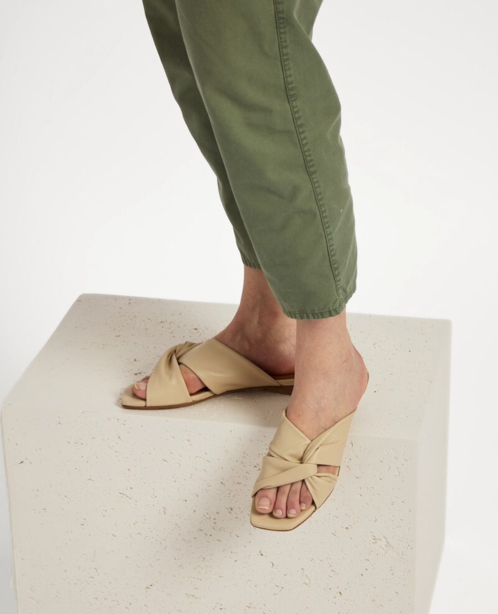 A pair of beige, open-toed sandals with a crisscross strap design, featuring a flat sole and a smooth, matte finish, worn by a person standing on a textured, off-white block, with green pants visible.