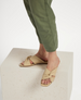 A pair of beige, open-toed sandals with a crisscross strap design, featuring a flat sole and a smooth, matte finish, worn by a person standing on a textured, off-white block, with green pants visible. 3