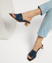 A pair of navy blue, open-toe mule sandals with a wide leather strap, square toe, and a low, light wooden block heel, worn by a person in light blue jeans. 3