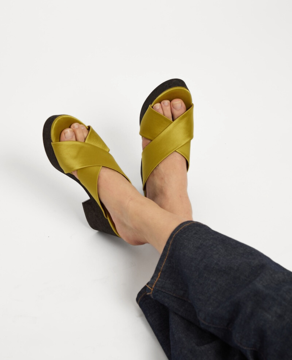 A pair of open-toed sandals with crisscrossing olive green satin straps and a dark cork wedge heel, worn by a person in dark denim pants.