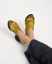 A pair of open-toed sandals with crisscrossing olive green satin straps and a dark cork wedge heel, worn by a person in dark denim pants. 2