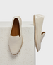 A pair of sleek, cream-colored slip-on shoes with a smooth leather texture, featuring a rounded toe, a low-profile rubber sole, and a subtle logo embossed on the insole. 1