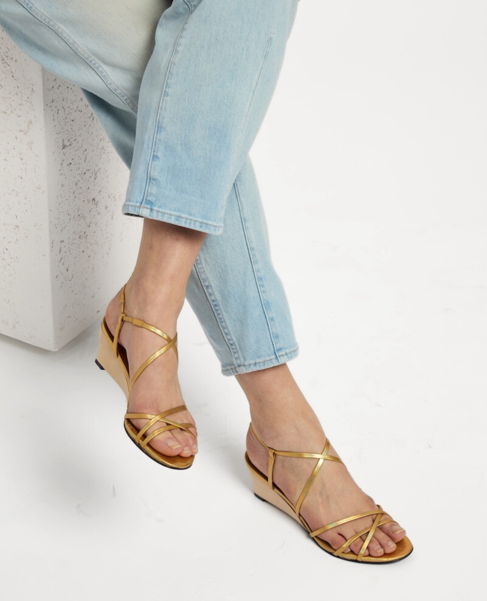 Golden strappy sandals with a low wedge heel, featuring a sleek, shiny finish and a minimalist design, worn with light blue jeans.