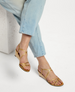 Golden strappy sandals with a low wedge heel, featuring a sleek, shiny finish and a minimalist design, worn with light blue jeans. 2