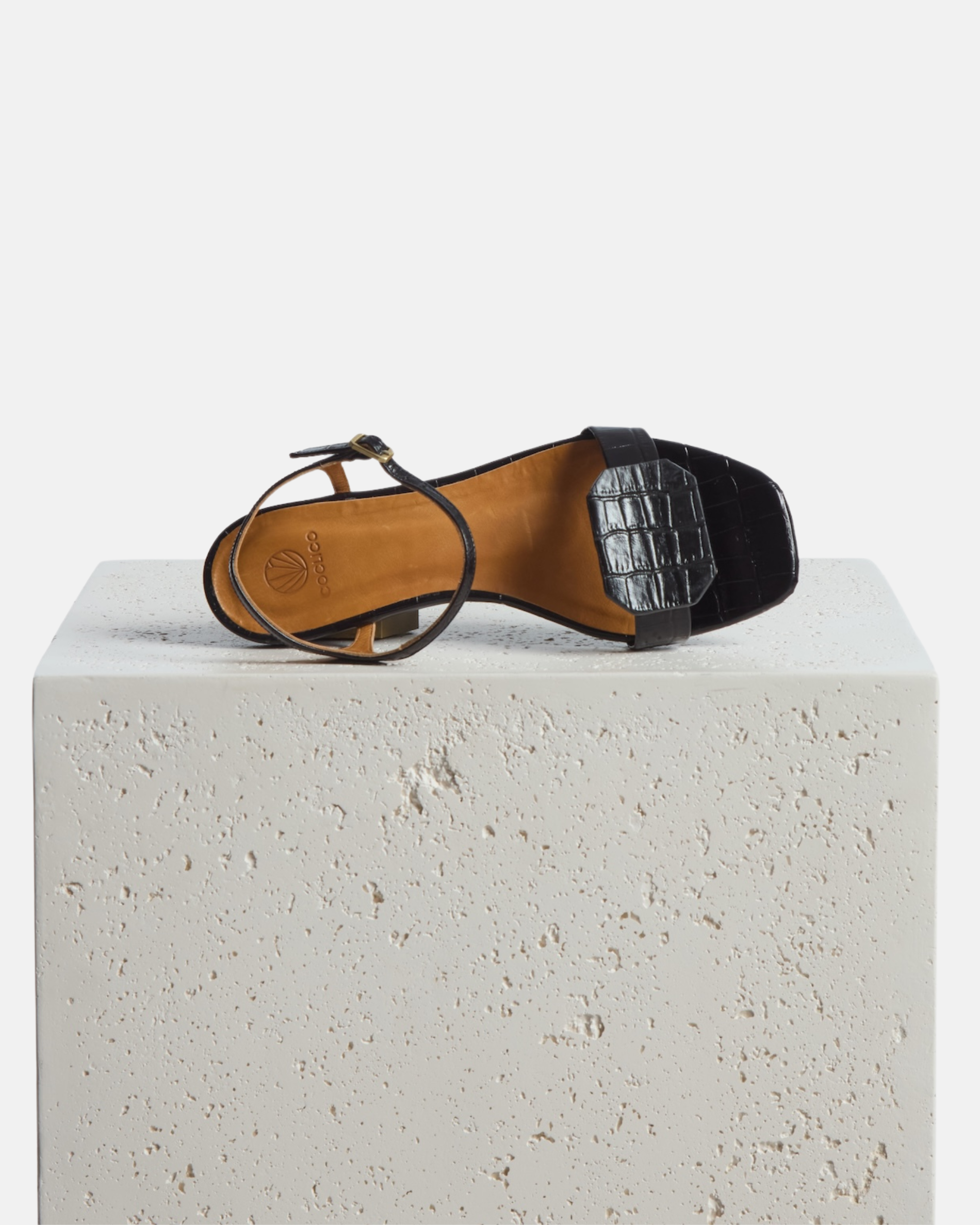 Shoe on a stone perdestal: black croc-embossed leather flat sandal with a slim ankle strap and a octagonal accent on the front strap, square toe, on a white background.