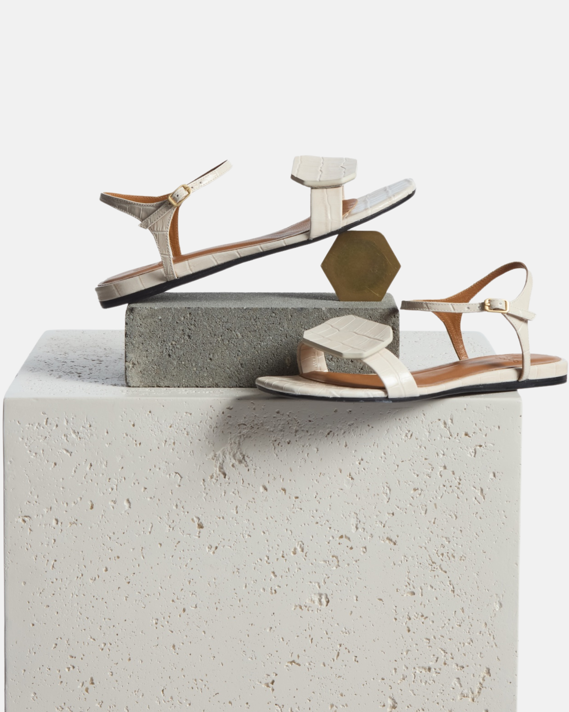 Shoe on plinth: Angled view of a bone-colored croc-embossed leather flat sandal with a slim ankle strap and a octagonal accent on the front strap, square toe, on a white background.