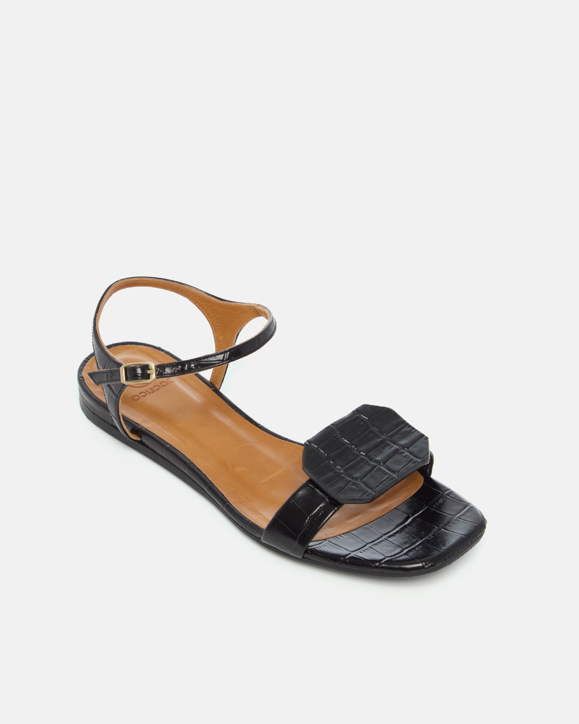Angled view of a black croc-embossed leather flat sandal with a slim ankle strap and a octagonal accent on the front strap, square toe, on a white background.