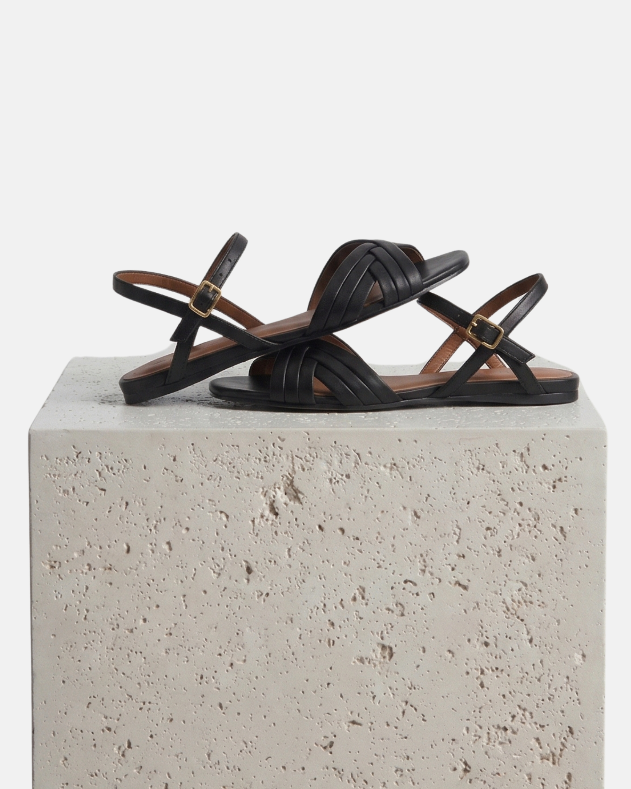 A black leather flat sandal with slim crisscross toe straps, buckle quarter strap, and square toe, artfully rranged on a white stone plinth.