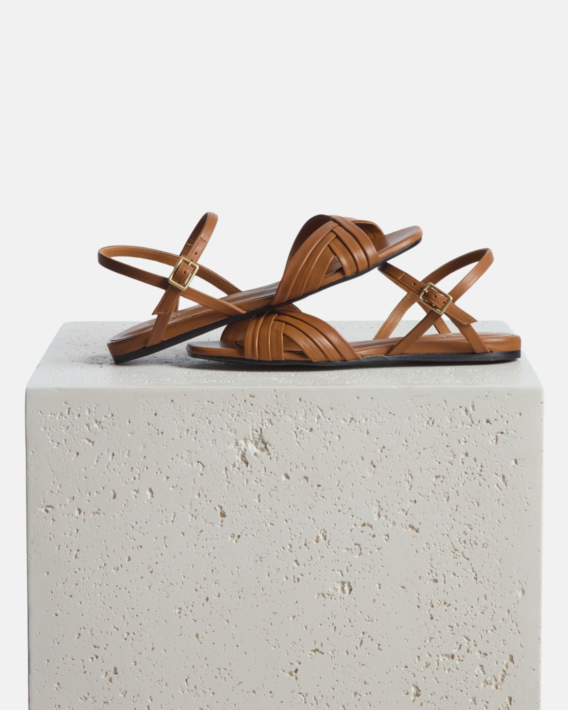 Sandles arranged on top of a pedestal: warm brown leather sandal with slim crisscross straps, buckle quarter strap, and a square toe on a grey background