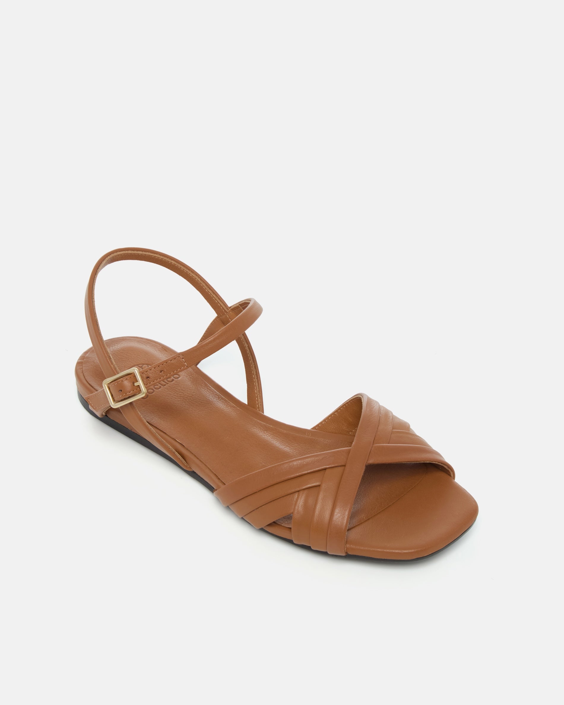 Angled view of a warm brown leather sandal with slim crisscross straps, buckle quarter strap, and a square toe on a grey background