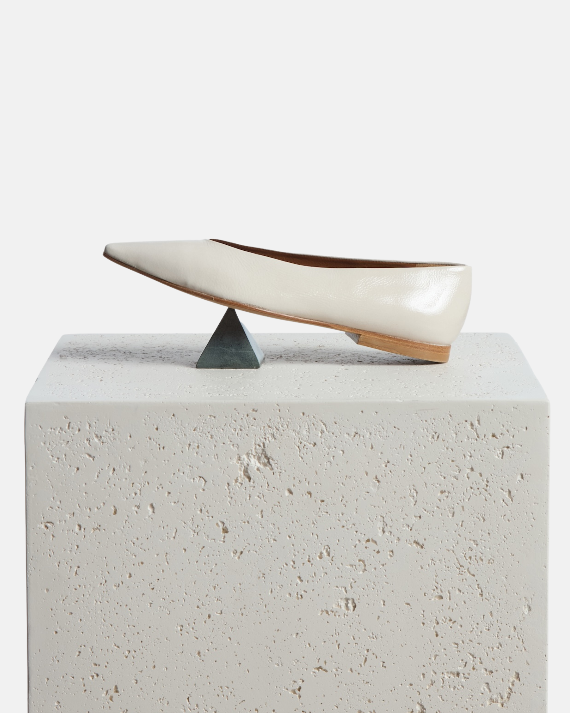 Shoe arranged artfully on a pedestal: off-white patent leather pointed-toe flat with clean lines and a flexible leather sole, on a white background.