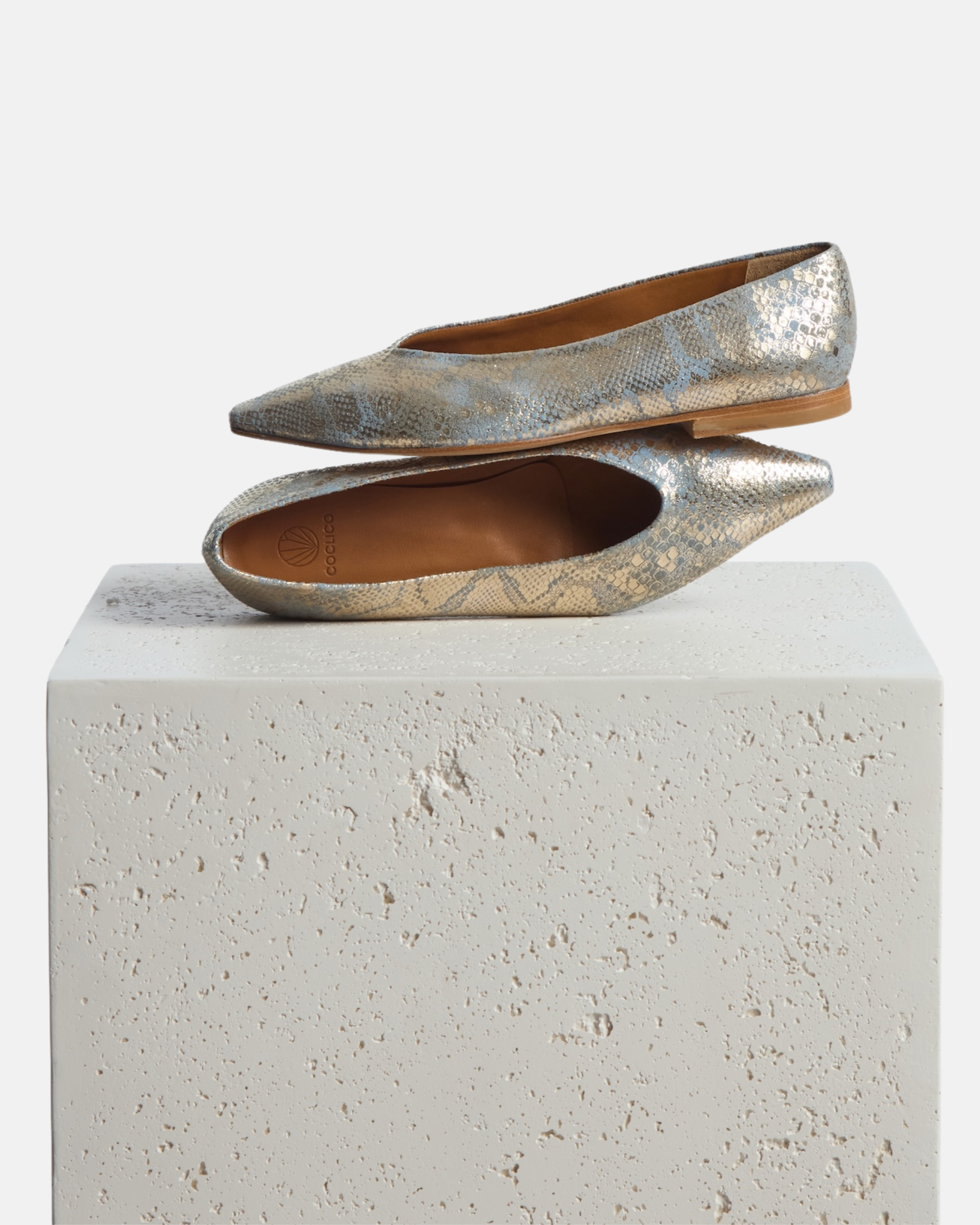 Shoes arranged on a pedestal: blue and gold metallic python print leather pointed-toe flat with clean lines and a flexible leather sole, on a white background.