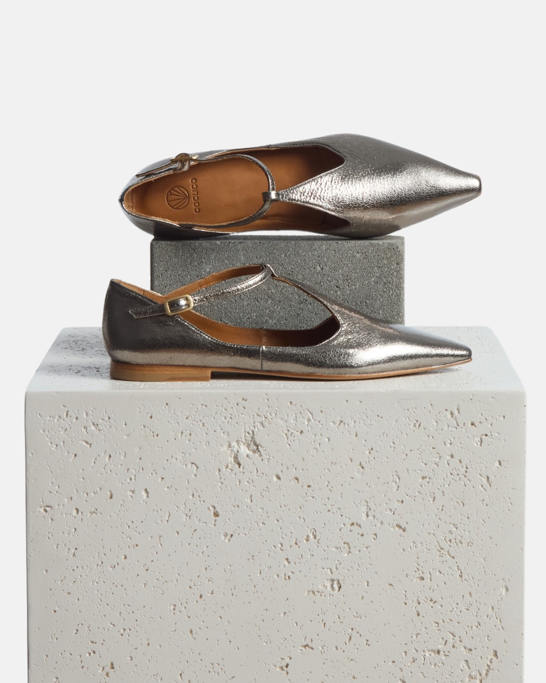 Shoes on a pedestal: pointed-toe silvery metallic T-strap flat with adjustable buckle and leather sole on a white background