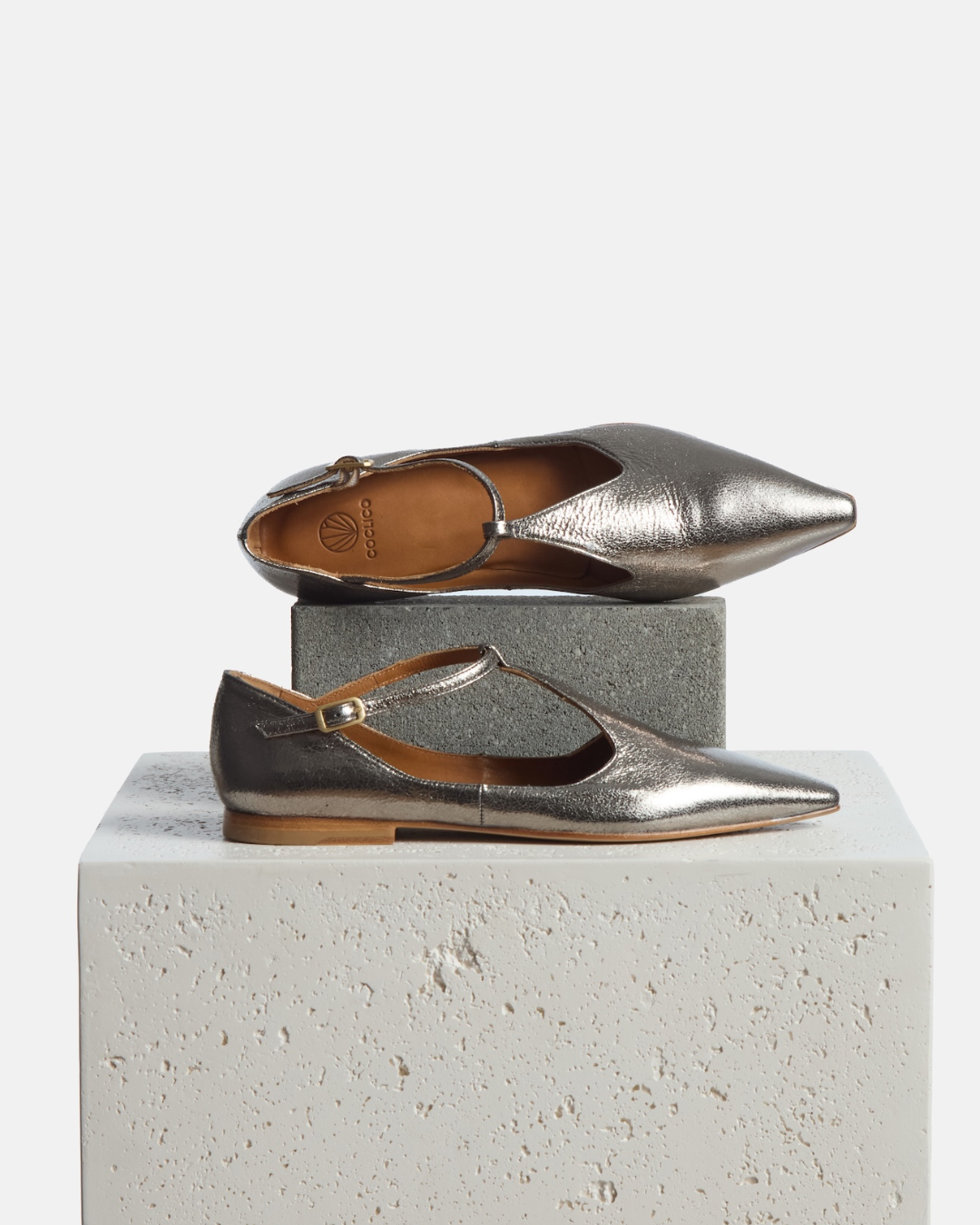 Shoes on a pedestal: pointed-toe silvery metallic T-strap flat with adjustable buckle and leather sole on a white background