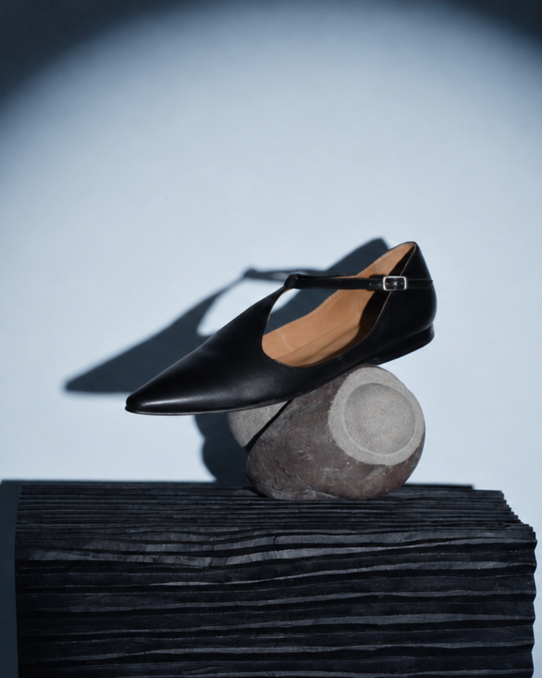 A single black pointed-toe Mary Jane flat with a tan interior and a small silver buckle, displayed on a round stone prop atop a dark textured pedestal against a cool light gray background.