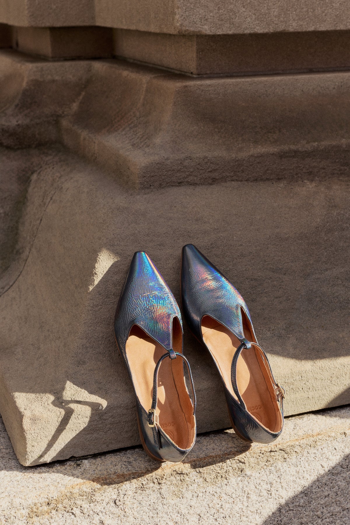The Coclico best selling Celeste flat in Carbon blue metallic rests on the side of a building's exterior