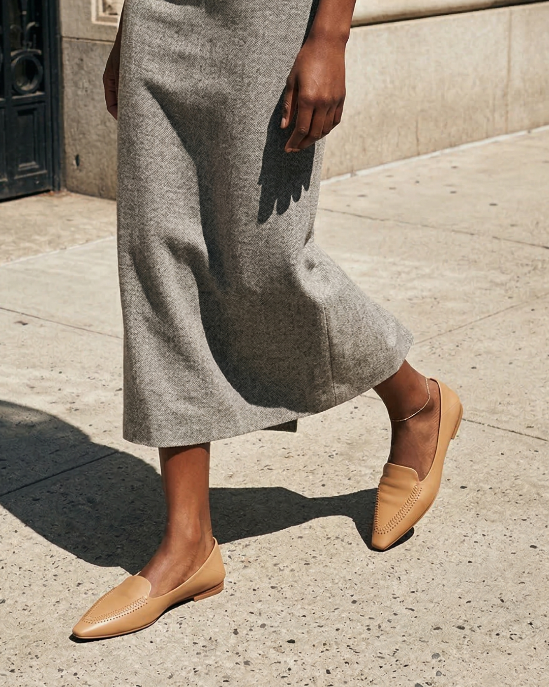 Woman walking on a city street in a long grey skirt and camel colored pointed-toe leather loafers.
