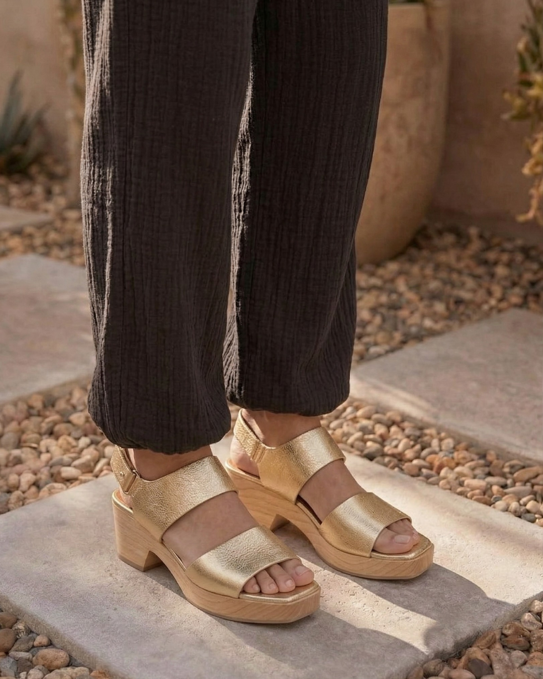 Legs of a woman wearing gold leather clog sandals and black cheesecloth pants standing in a sunny courtyard.