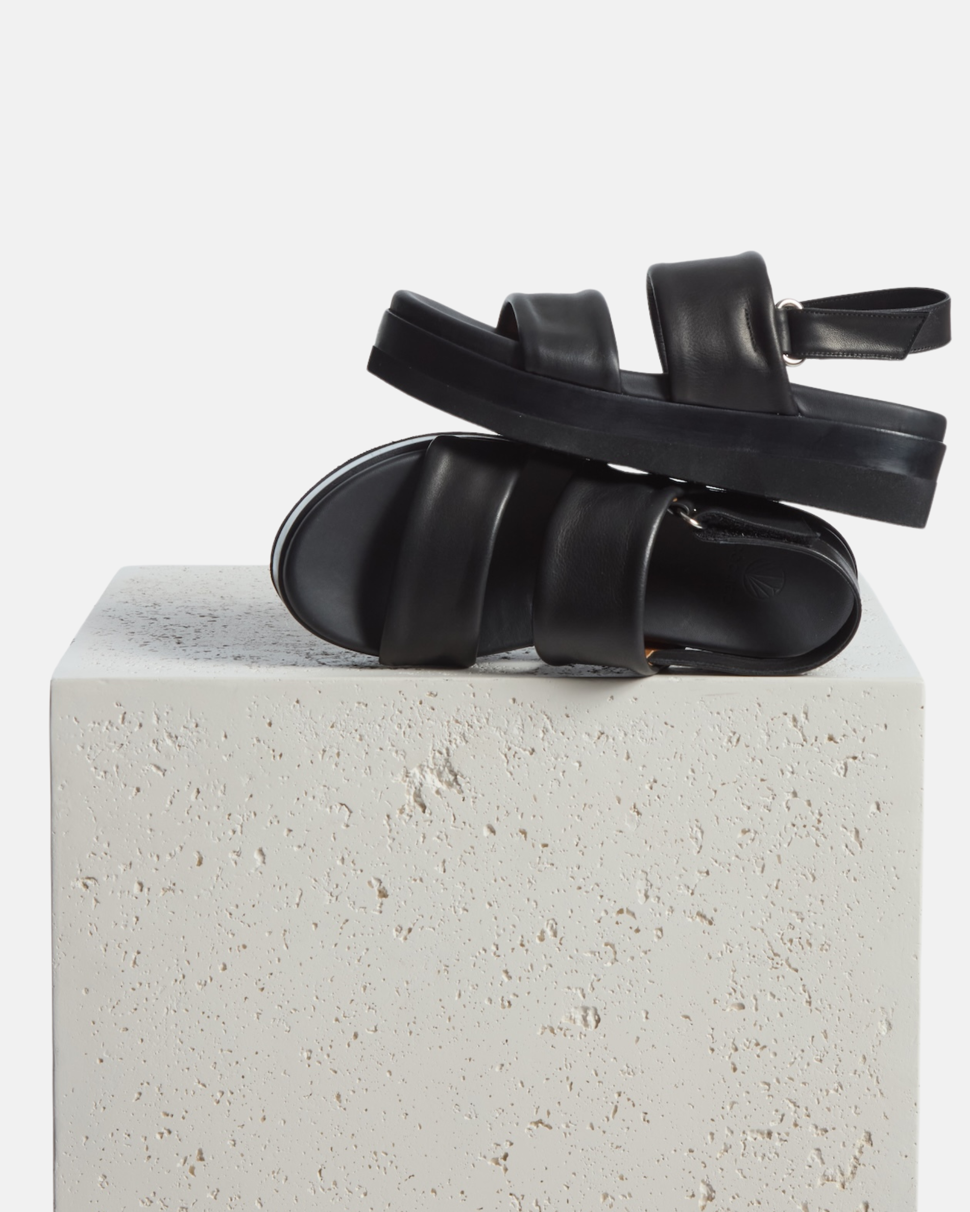 A sleek black sandal with a thick EVA sole, featuring two wide leather straps across the foot and an adjustable ankle strap, resting on a textured white surface.