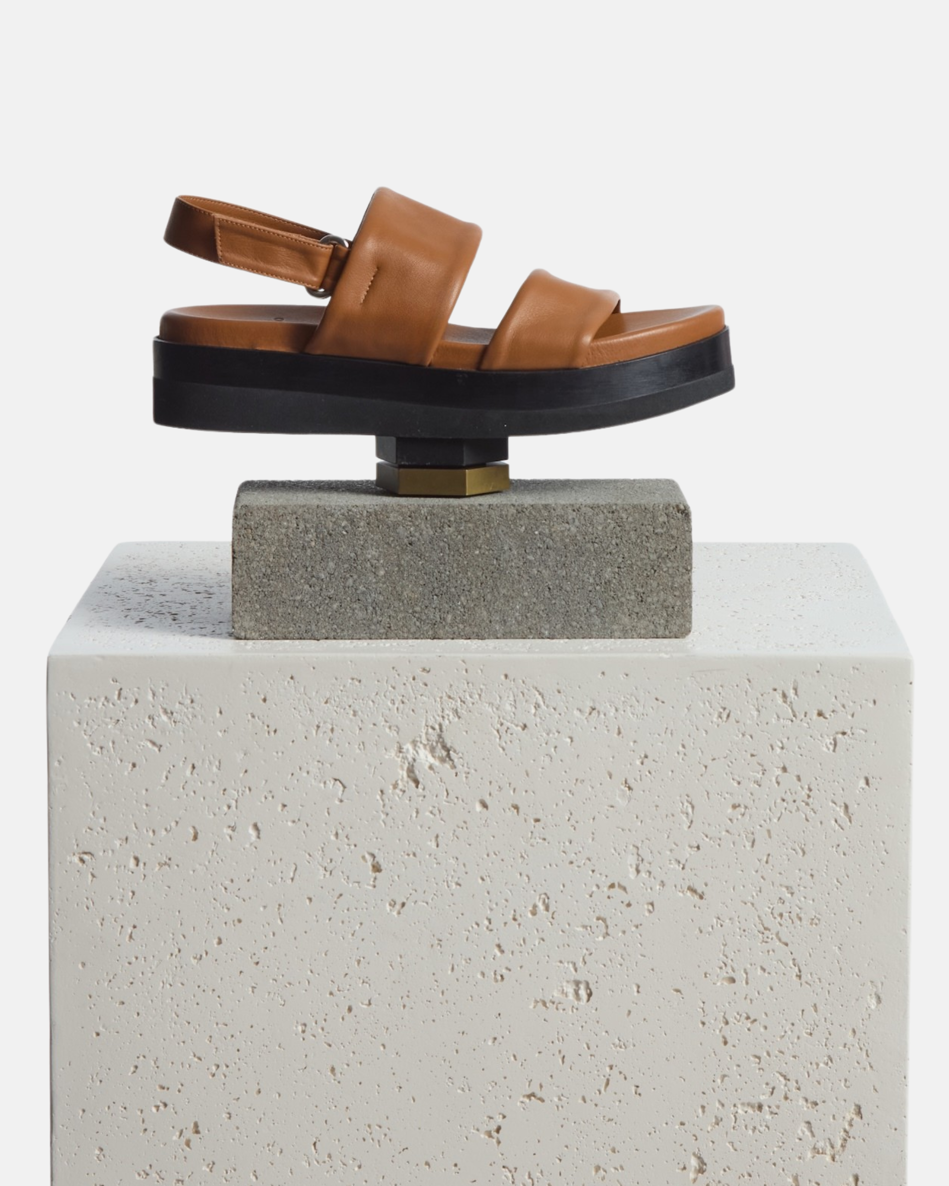 Shoe artfully arranged on a plinth: brown leather wedge sandal with wide tubular straps, velcro slingback, and a lightweight EVA platform sole
