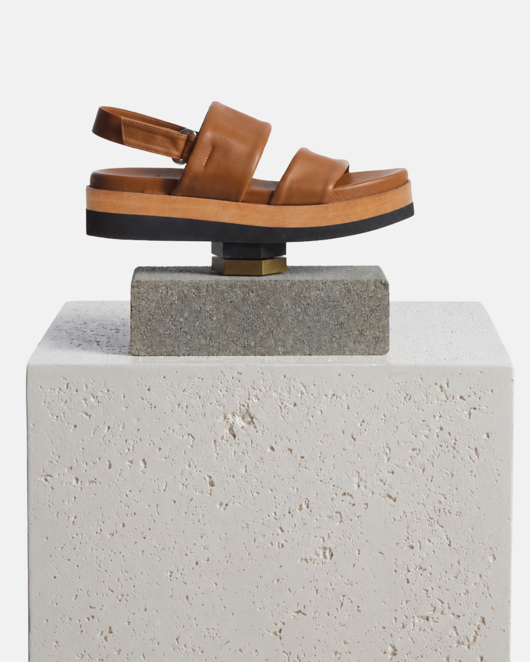 Shoe artfully arranged on a plinth: brown leather wedge sandal with wide tubular straps, velcro slingback, and a lightweight EVA platform sole