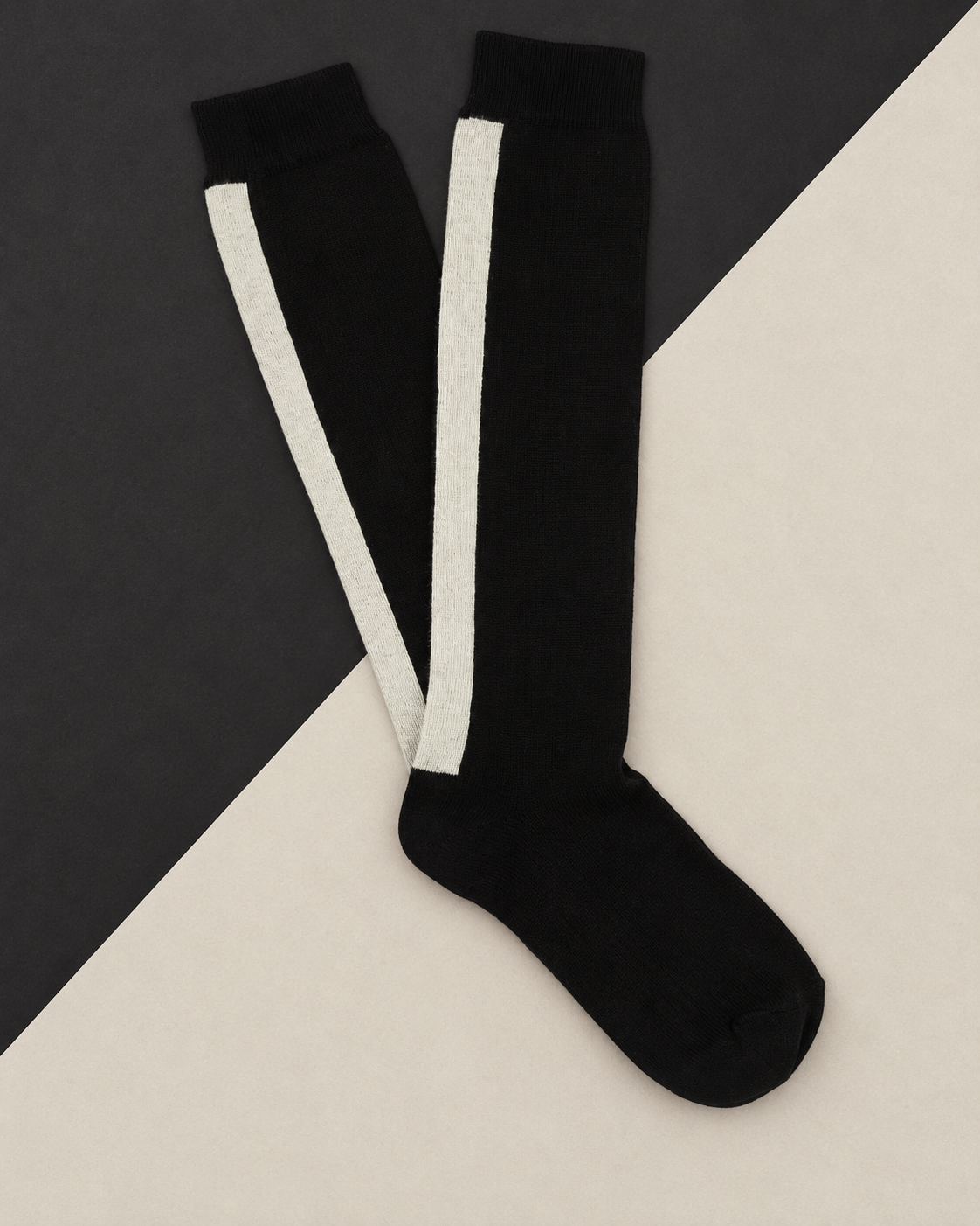 Escuyer knee high socks in Black with an ecru side stripe, laid flat on a two-tone black and cream background