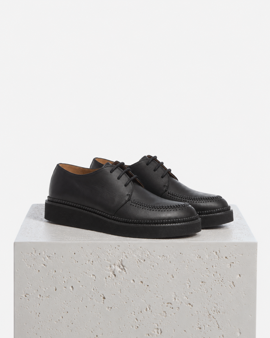A pair of black leather lace-up shoes with brogue detailing and chunky black soles, displayed on a textured white stone pedestal against a clean white background.