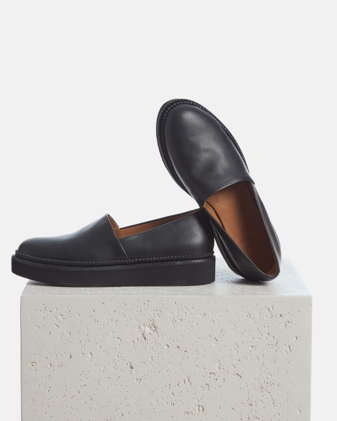 Coclico Ginsberg Loafer in black leather, flatform slip ons with apron fronts styled on a stone plinth