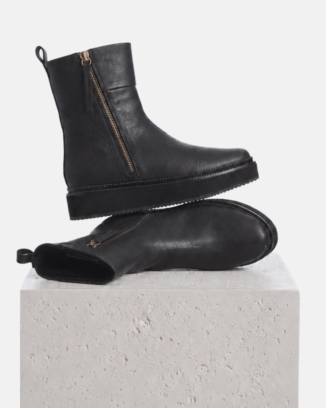 A pair of black leather, shearling lined ankle boots with side zippers, and a wedge sole, displayed on a textured white stone block against a light gray background.