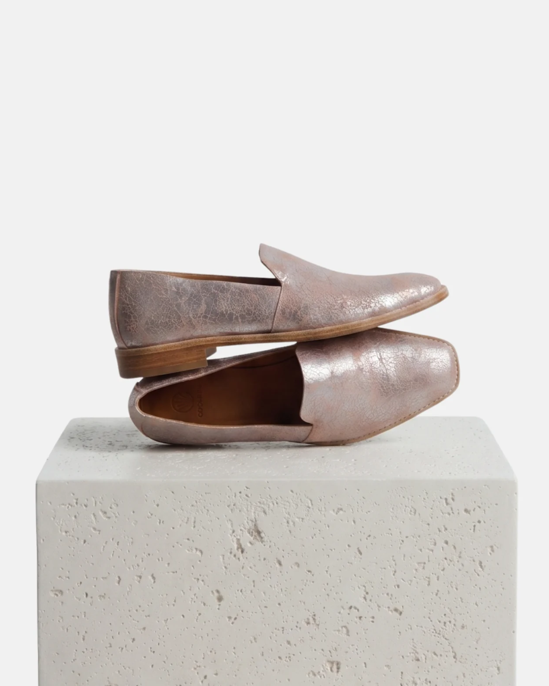 A pair of metallic rose gold leather loafers with a distressed finish, displayed on a textured light gray concrete block against a clean grey background.