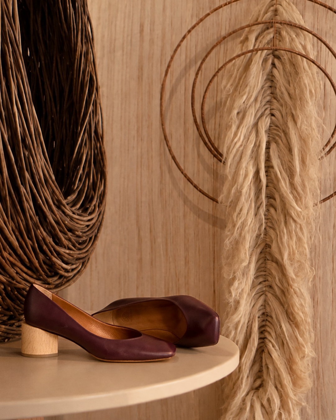 A pair of women's mid-heeled pumps in deep burgundy leather with light wood block heels, styled on a light beige table against a warm wood-paneled wall with decorative dried grass elements.