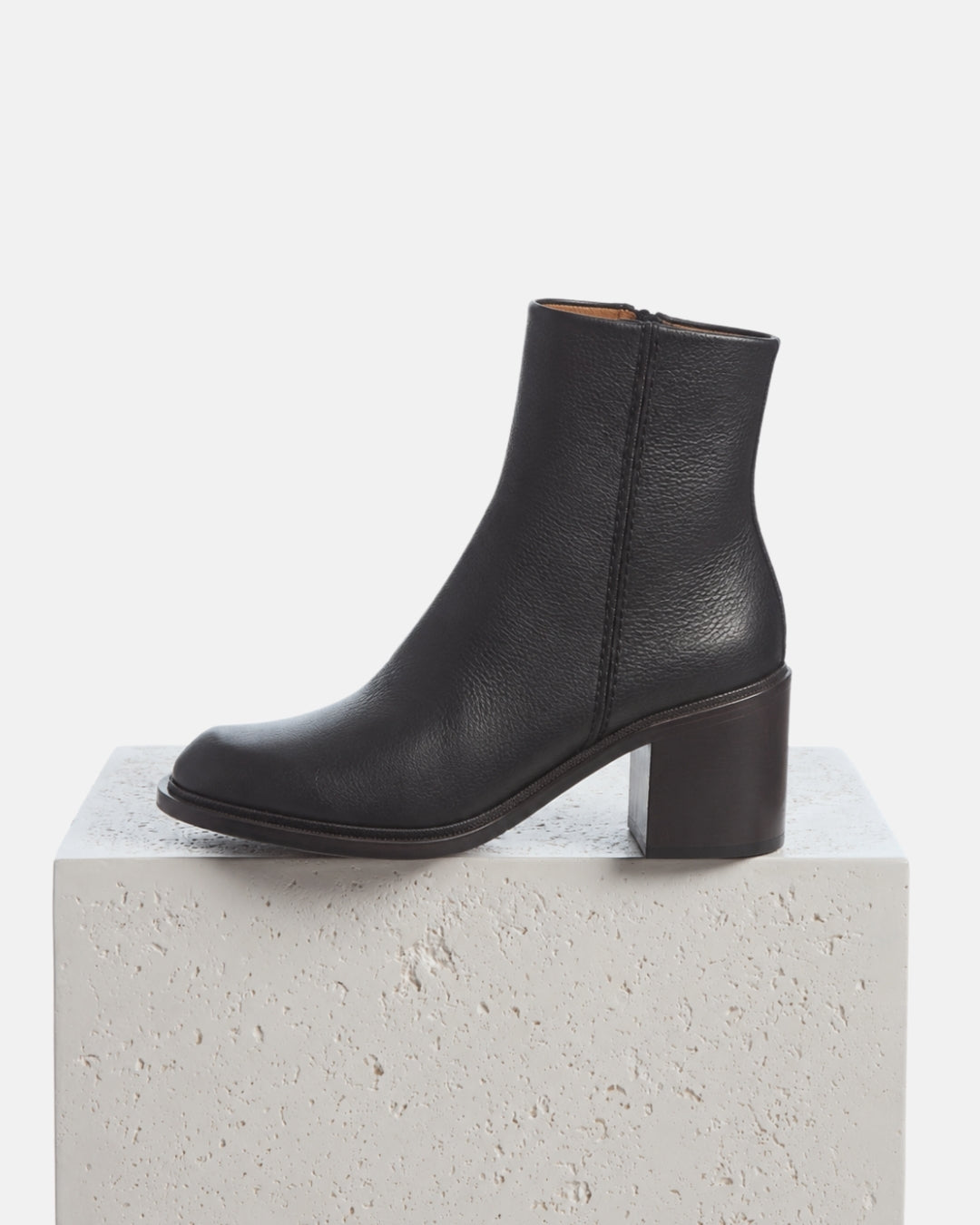 A black leather ankle boot with a block heel and butted side seam, displayed n a textured white stone pedestal against a clean light grey background.