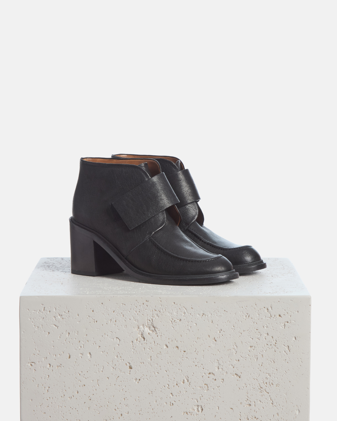 Coclico Lilya Boot in black leather, crossover strap ankle boots with block heels styled on a stone plinth