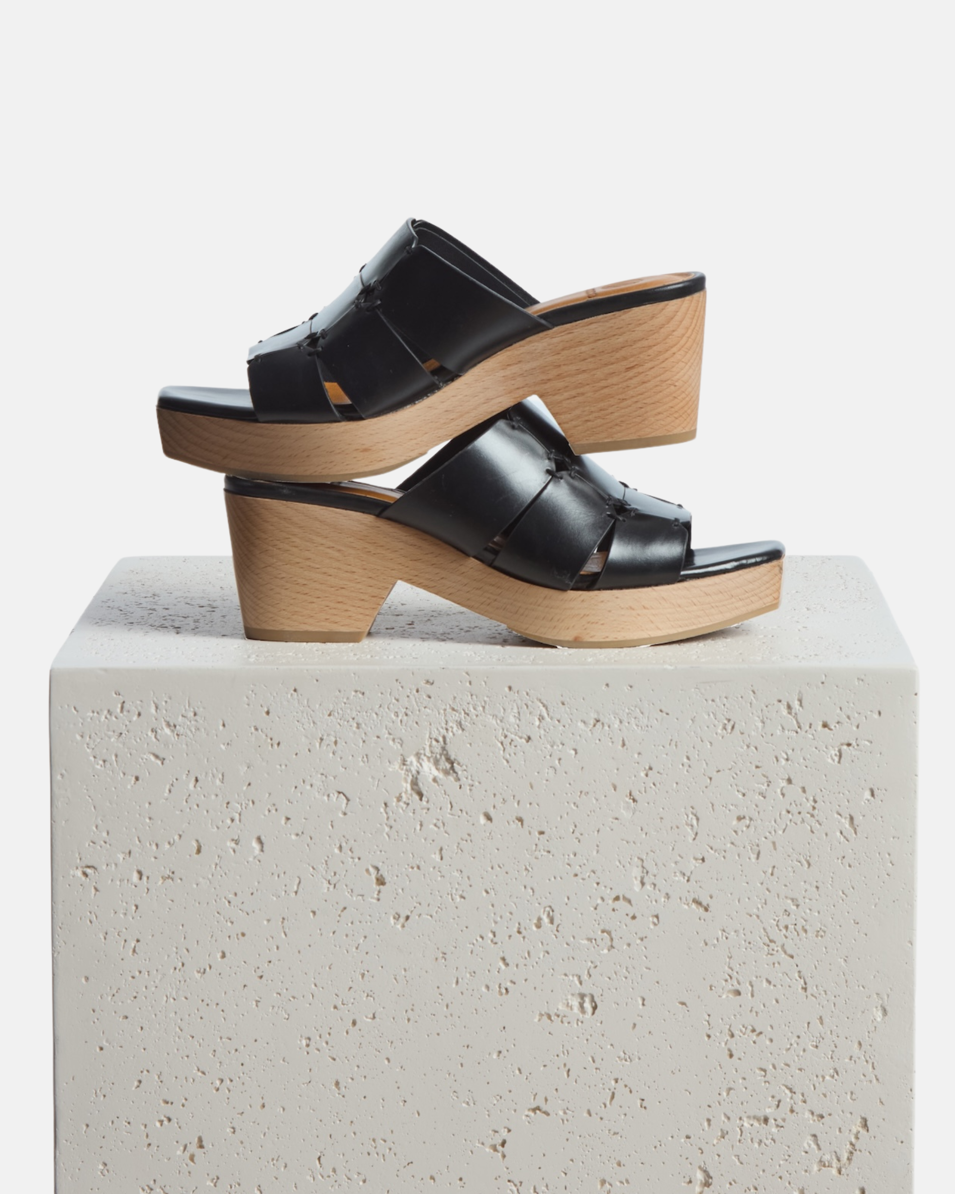 Clogs stacked on a plinth: clog sandal in black leather featuring linked straps, wooden heel, and open toe slip-on design
