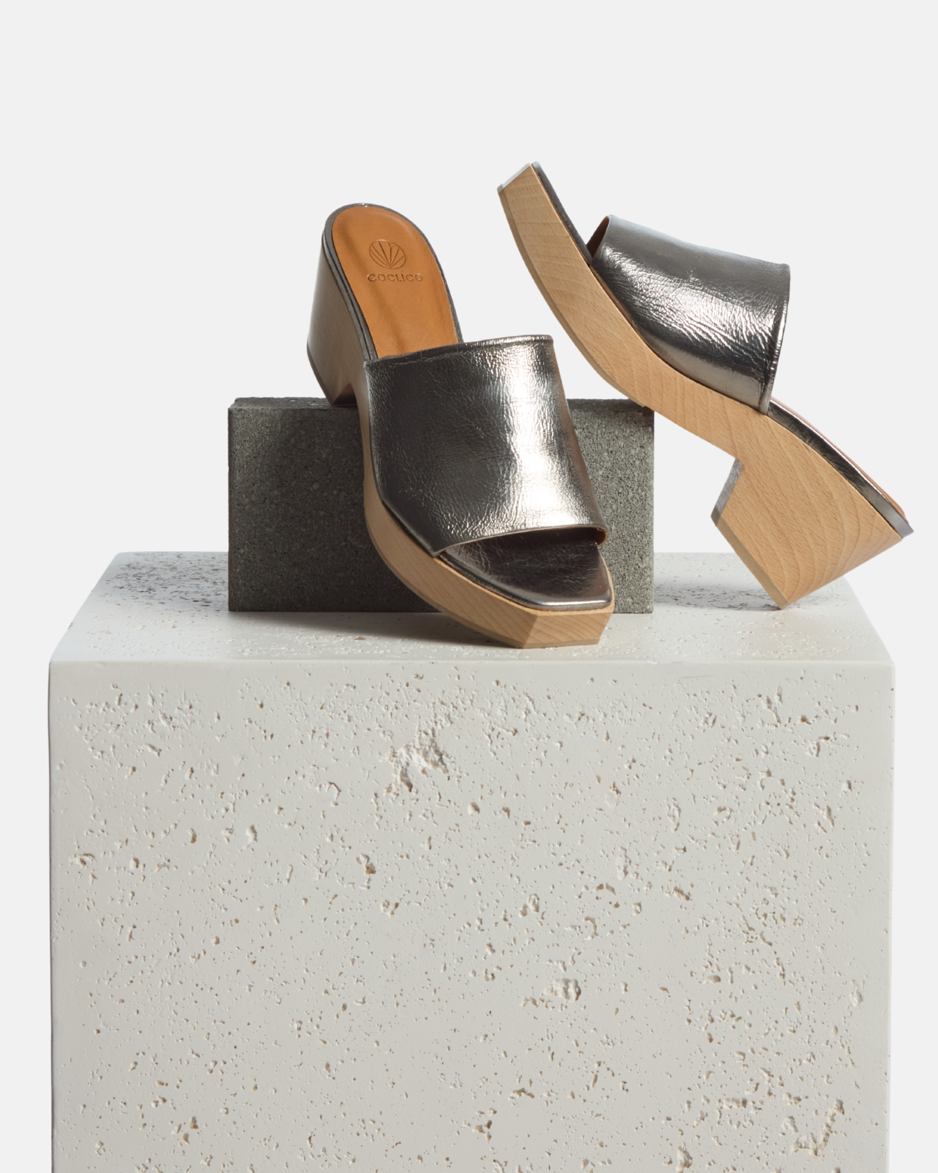 A pair of open-toe mule clog sandals featuring a metallic silver leather upper with a textured finish, set on a light wooden block heel and platform with a smooth surface.